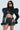 Wearing the ultra-cropped KEEP IT COMING TOP with structured, puffed shoulders and black leather shorts, a person with long hair, sunglasses, and multiple rings poses against a plain white background.