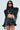 A person in the KEEP IT COMING TOP showcases a shiny, leather-like quilted crop top with exaggerated shoulders and matching shorts, boasting an ultra-cropped fit. They wear sunglasses and gold rings, complementing their long dark hair.