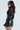 Modeling the KEEP IT COMING TOP in glossy black with ribbed texture, ultra-cropped fit, and an open back elegantly tied with strings, they pair it with black leather shorts and sunglasses, their long dark hair cascading against a plain light gray background.