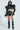 A woman exudes confidence in her Keep It Coming Top, a futuristic black leather outfit with paneled puffer shoulders and an ultra-cropped fit. She pairs it with matching high-waisted shorts, knee-high boots, sleek black sunglasses, and sports long dark hair.