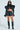 A woman confidently wears a futuristic black KEEP IT COMING TOP with exaggerated shoulders and long sleeves. Her ultra-cropped style is paired seamlessly with high-waisted shorts, knee-high boots, and sunglasses against a plain background.