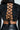 A person wears the KEEP IT COMING TOP, a glossy black ruched top with an open back and crisscross laces. It's paired with black leather pants on a light gray backdrop, creating a striking look.