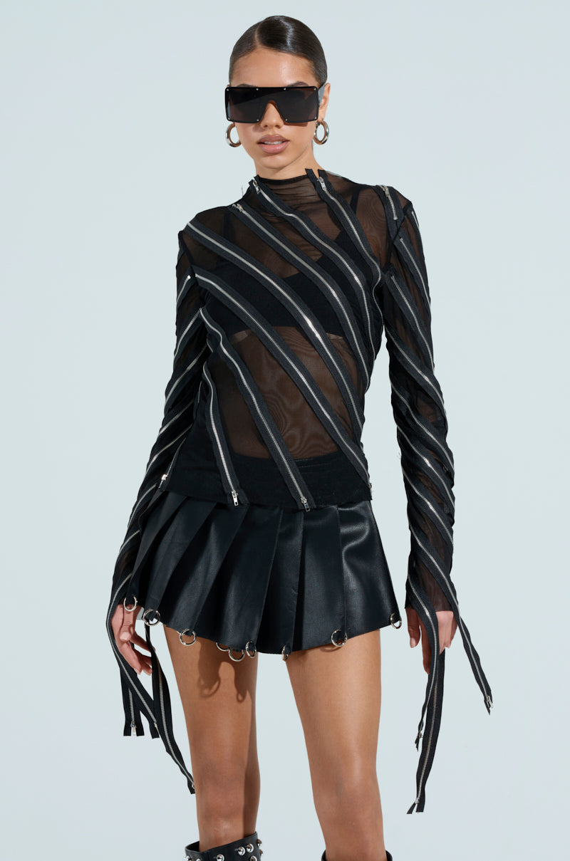 Wearing the CROSS ZIP MESH TOP, a fitted black outfit with zipper panels and a pleated mini skirt, the person pairs it with dark sunglasses and slicked-back hair, exuding confidence against a plain backdrop.