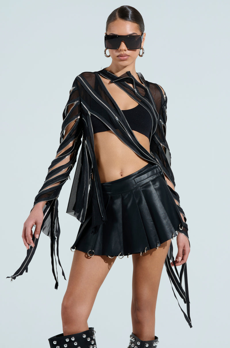 A person poses in a stylish CROSS ZIP MESH TOP featuring cutout sleeves, zipper panels, and a fitted silhouette, paired with a pleated black skirt and knee-high boots with silver studs. They wear large sunglasses and hoop earrings, with hair tied back.