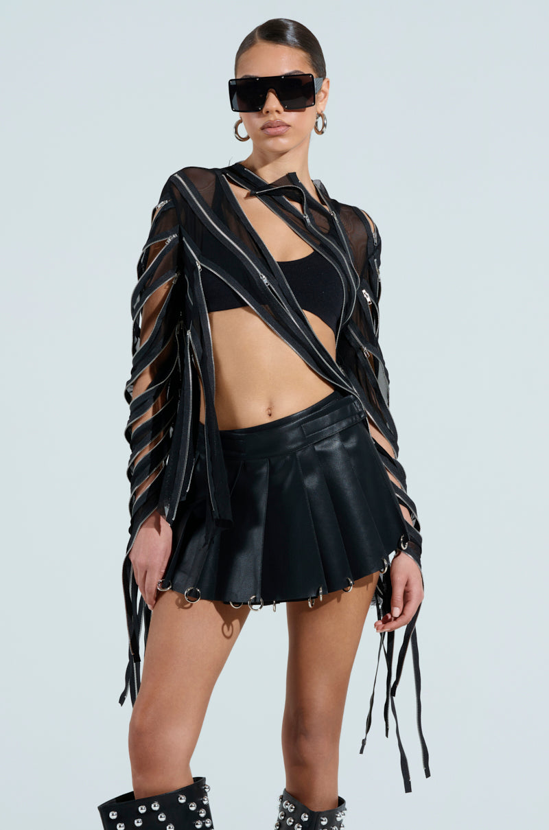 Wearing oversized black sunglasses and a CROSS ZIP MESH TOP, paired with a mesh jacket with long strips, a pleated black leather skirt with silver details, and knee-high studded boots, the person poses confidently against a plain background.