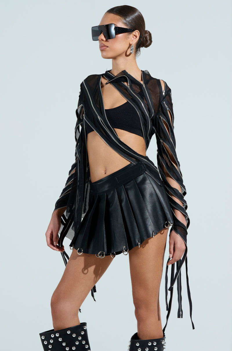A fashion-forward woman poses confidently in a black outfit featuring a CROSS ZIP MESH TOP, shredded leather jacket with zipper panels, pleated leather skirt, and knee-high boots with metal studs. Oversized sunglasses and braided hair add a futuristic touch.