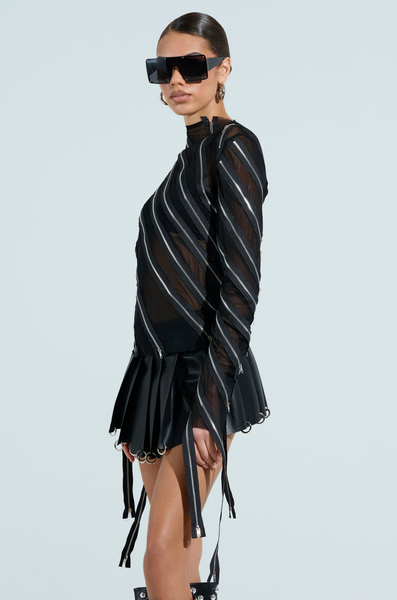 Dressed in a chic black ensemble with diagonal silver stripes, they wear a CROSS ZIP MESH TOP and pleated skirt. With short hair, large black sunglasses, and boots, they stand against a light background.