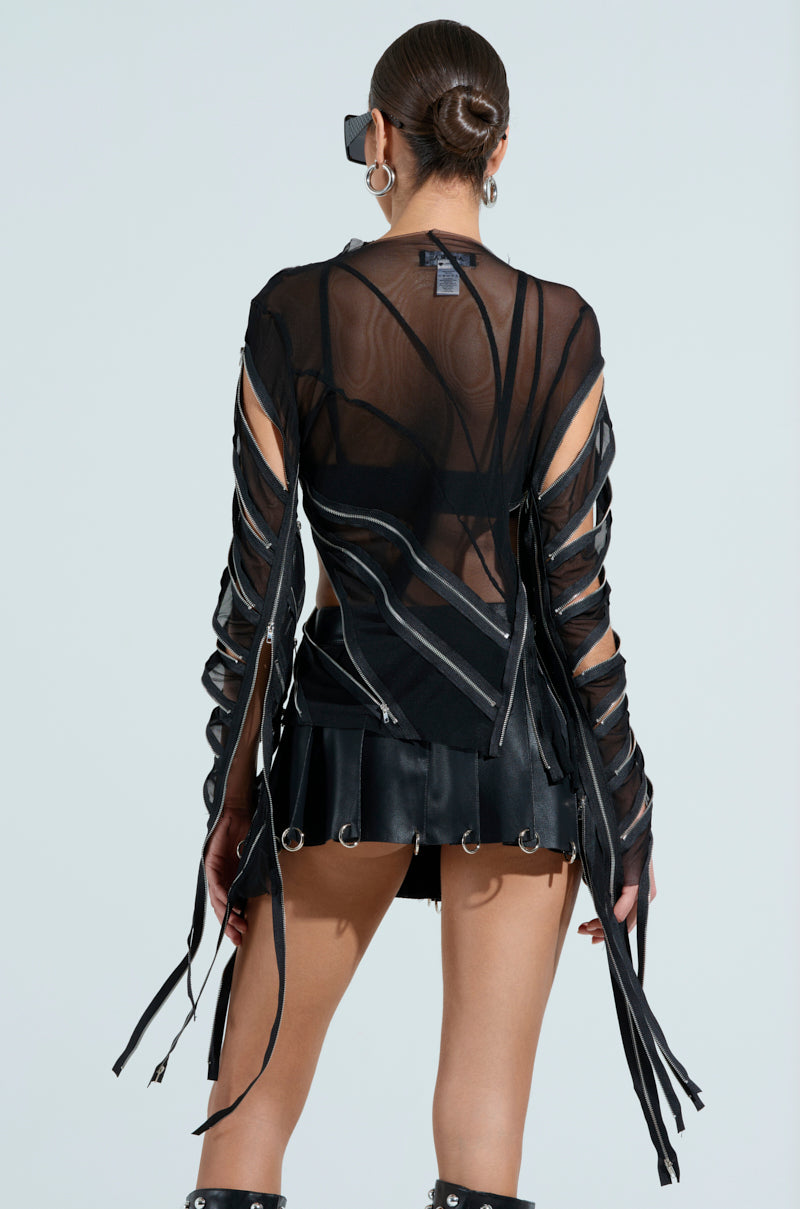 A person with hair in a sleek bun wears sunglasses and a fitted silhouette featuring the CROSS ZIP MESH TOP, highlighted by zipper panels, cutouts, and a black leather skirt adorned with metal rings. The outfit has long, ribbon-like extensions against a plain light gray background.
