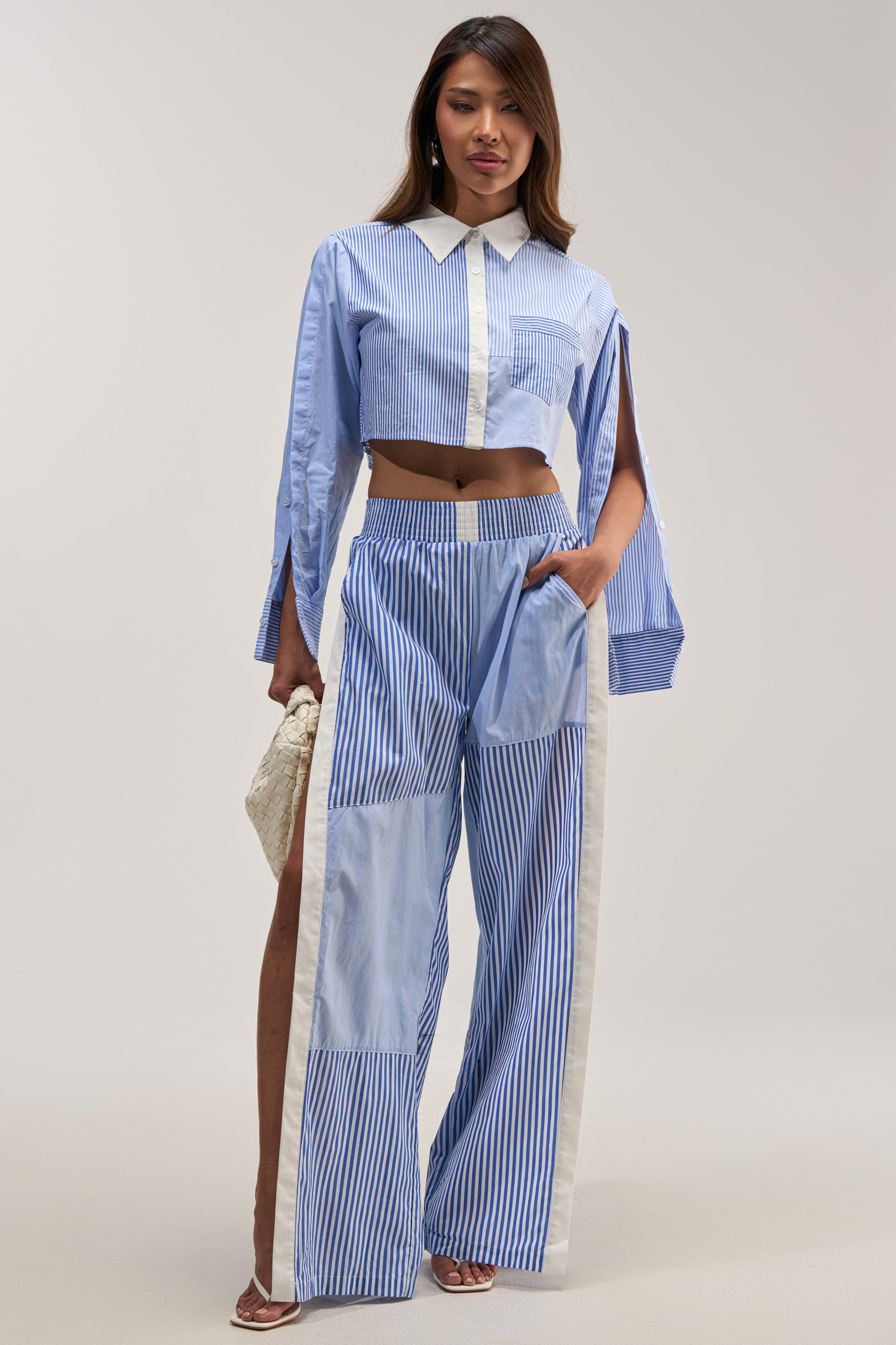 A model poses in a studio wearing the REESE MIXED STRIPE POPLIN WIDE LEG PANT, featuring blue and white vertical stripes and side detailing. She styles the high-rise, wide-leg pants with a matching long-sleeved, cropped button-up shirt—creating a chic look perfect for summer casual wear. Accessorized with a small woven white handbag and strappy heels.