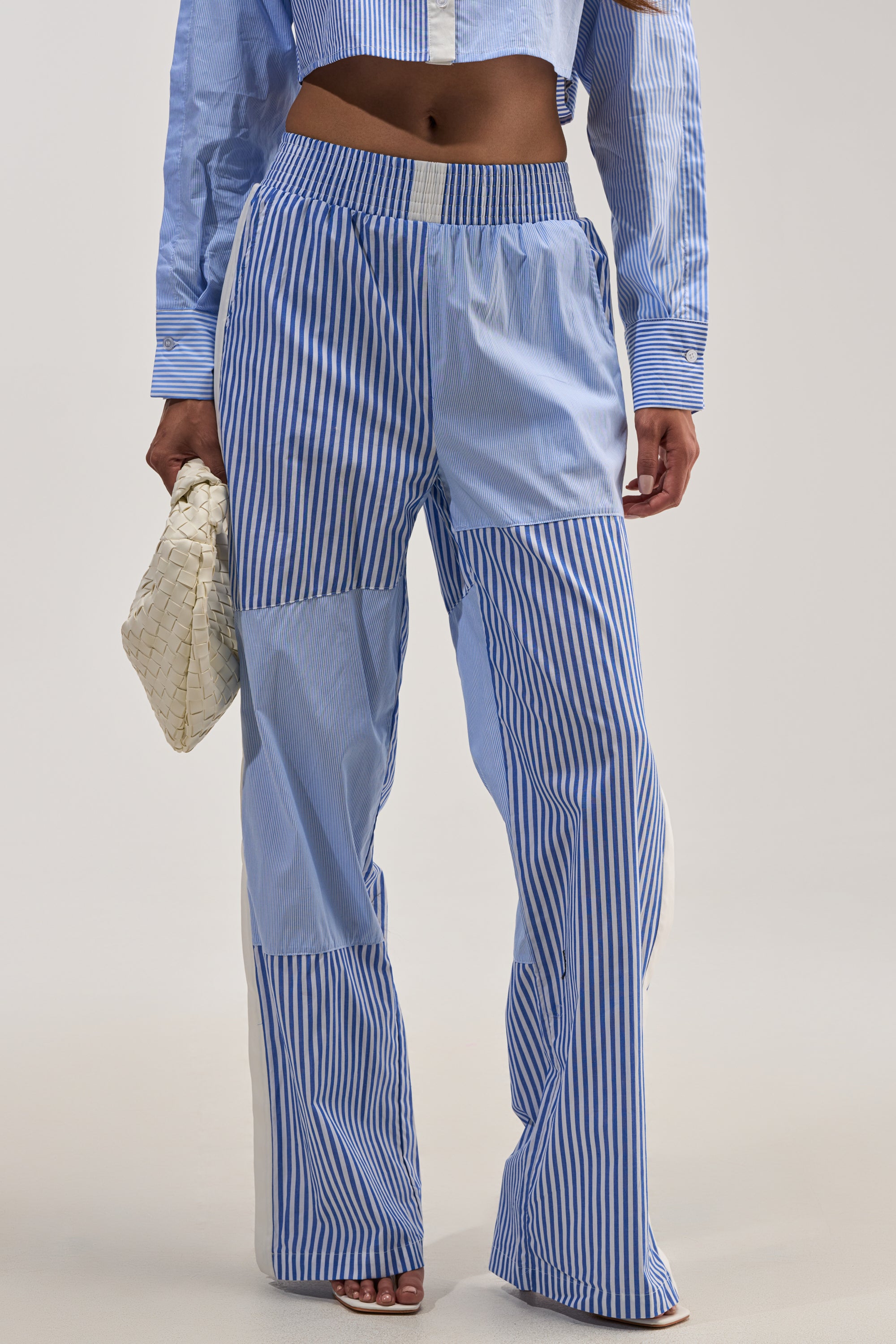 A person wearing the REESE MIXED STRIPE POPLIN WIDE LEG PANT with blue and white vertical stripes, paired with a matching cropped button-down shirt and holding a woven white clutch bag; only the lower body is visible.