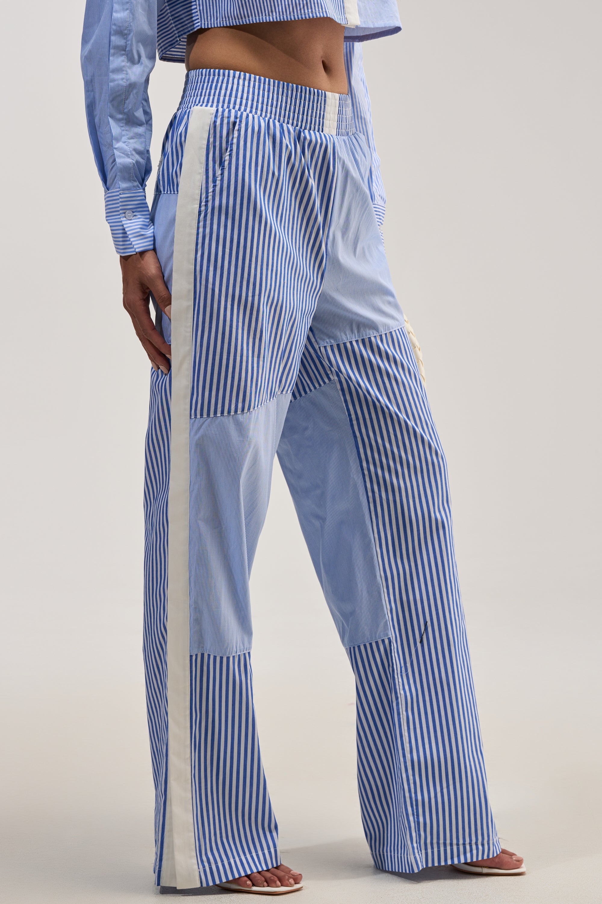Wearing the REESE MIXED STRIPE POPLIN WIDE LEG PANT featuring loose, blue and white vertical stripes with white side stripes, styled with a matching cropped shirt. The high-rise, wide-leg silhouette offers a relaxed look ideal for summer casual wear. The image shows the lower body and midriff against a plain, light background.