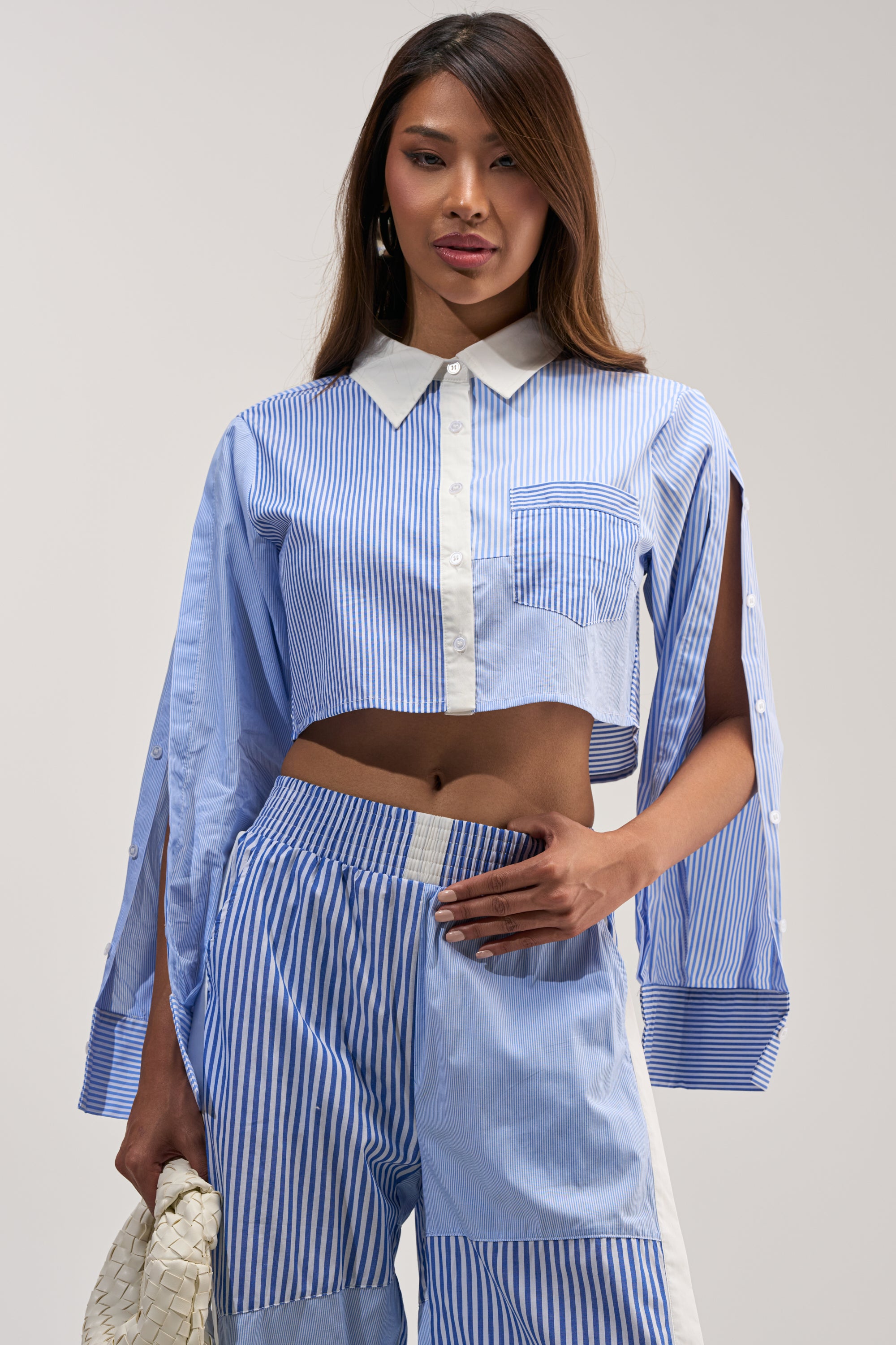 A woman poses confidently against a neutral background, wearing the REESE MIXED STRIPE POPLIN BUTTON DOWN BLOUSE—an asymmetrical, blue and white striped cropped style—paired with matching pants and holding a white textured clutch bag.