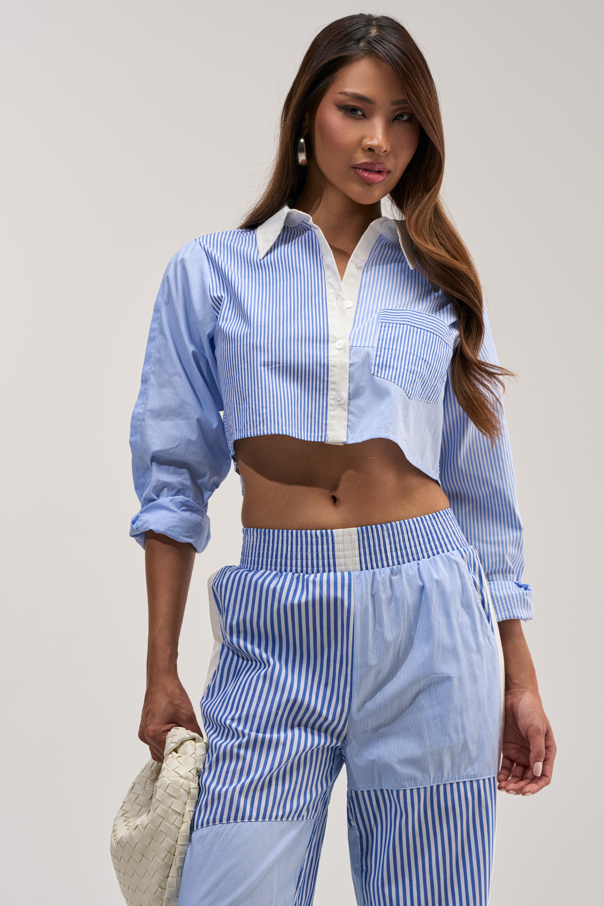 Wearing the REESE MIXED STRIPE POPLIN BUTTON DOWN BLOUSE, styled as a cropped blue and white striped shirt with matching high-waisted pants, she holds a textured cream clutch while standing against a plain background.