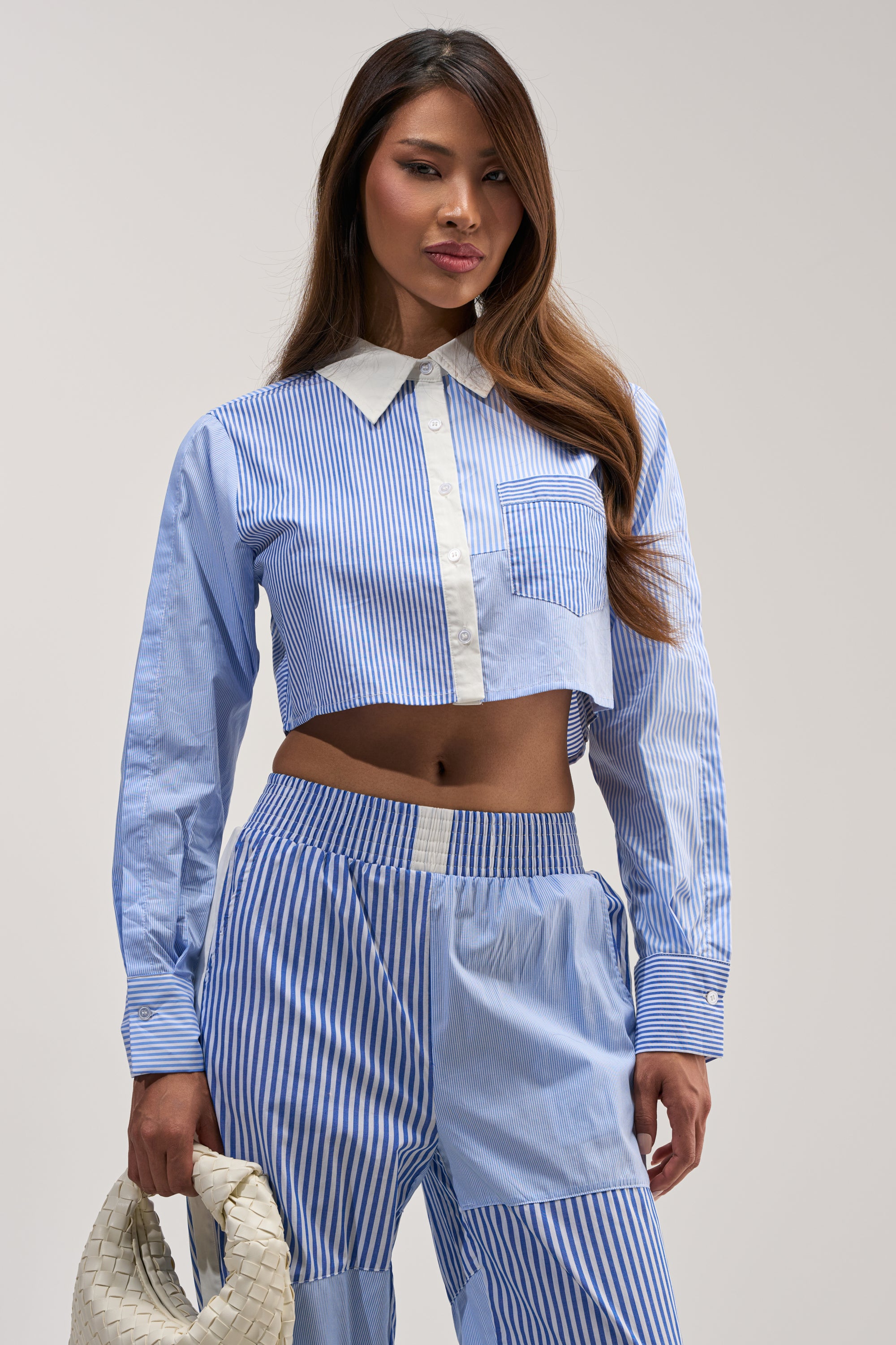 A woman wearing the REESE MIXED STRIPE POPLIN BUTTON DOWN BLOUSE with matching high-waisted pants holds a white woven bag, standing against a plain light background.