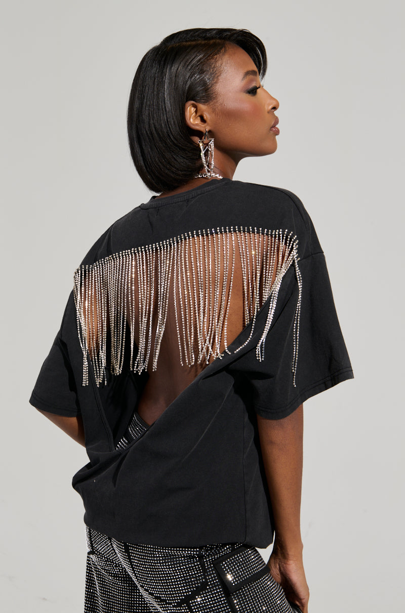 A woman showcases her fashion-forward style in the Smoke & Mirrors Rhinestone Fringe Mineral Wash T-Shirt, featuring an open back and chic rhinestone detailing. She accessorizes with statement earrings and patterned pants, while her sleek bob stands out against the neutral background. 0