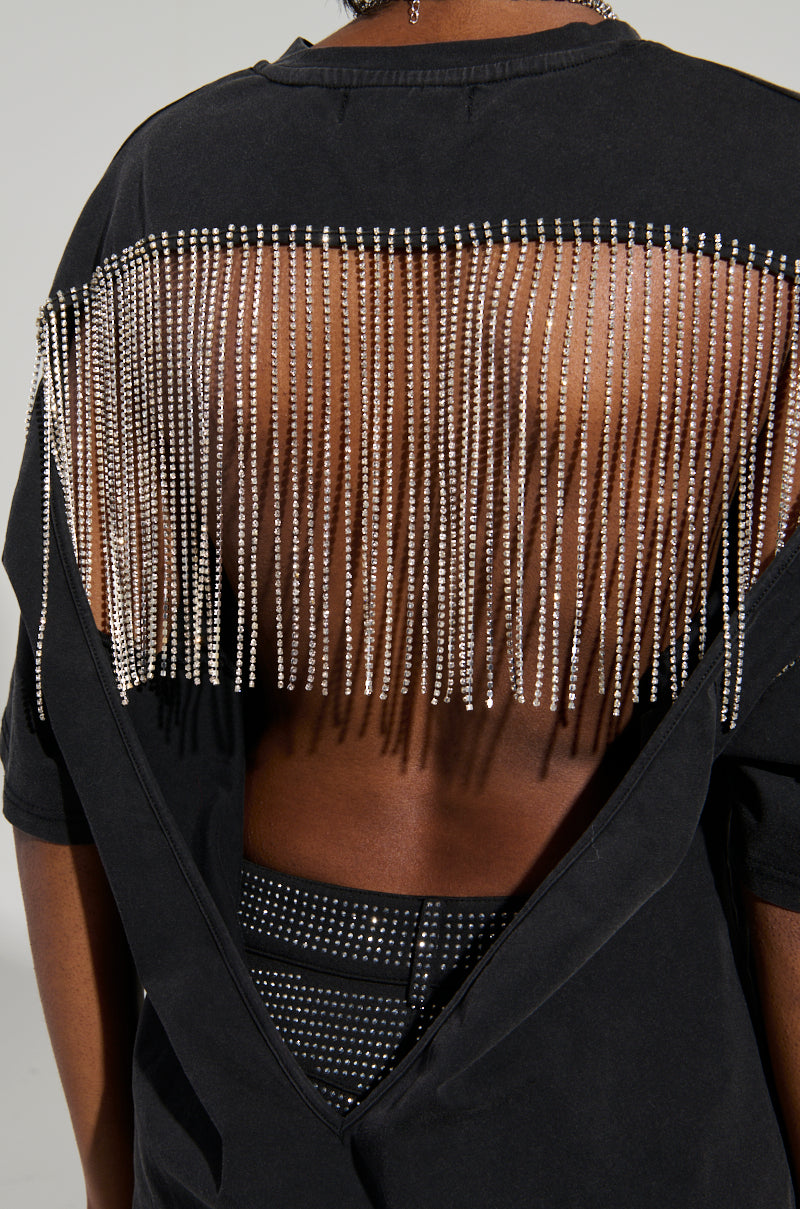 A person is wearing the SMOKE & MIRRORS RHINESTONE FRINGE MINERAL WASH T-SHIRT, which features a black finish and a cut-out back adorned with rhinestone fringe. This top is paired with dark, studded pants, and the image highlights the intricate details of this super stretchy outfit. 9
