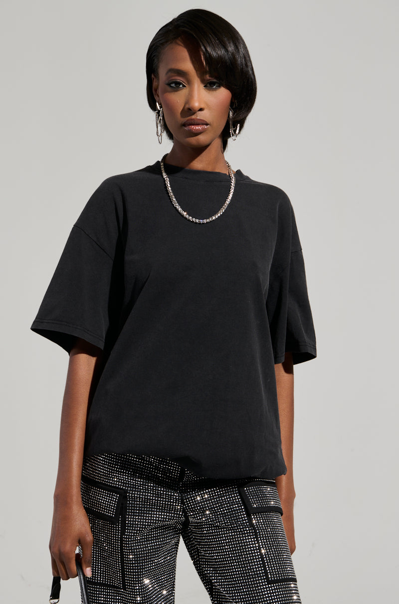 A person with short dark hair wears the SMOKE & MIRRORS RHINESTONE FRINGE MINERAL WASH T-SHIRT, perfectly paired with shiny, patterned pants. Rhinestone fringe enhances their ensemble, complemented by a chain necklace for added flair. They hold a small item in their hand, and the plain backdrop highlights their striking style. 1