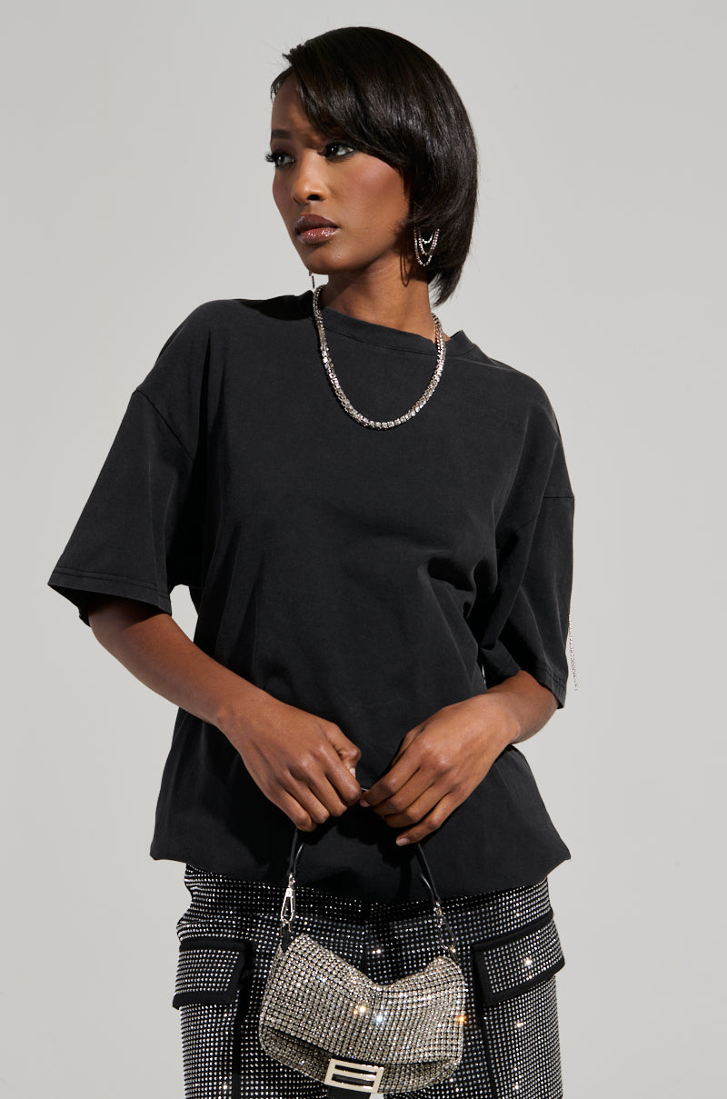 A person wearing a SMOKE & MIRRORS Rhinestone Fringe Mineral Wash T-Shirt paired with checkered pants and accessorized with a rhinestone fringe necklace poses against a plain background, holding a small, silver, textured handbag. 2