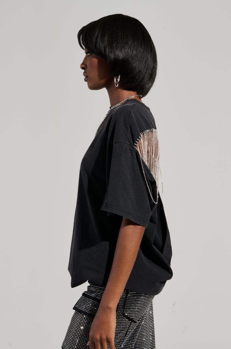A person with short, dark hair is wearing a SMOKE & MIRRORS RHINESTONE FRINGE MINERAL WASH T-SHIRT in black, featuring rhinestone fringe on one shoulder, paired with sparkling pants. They are standing in profile, looking to the left against a plain and light-colored background. 3