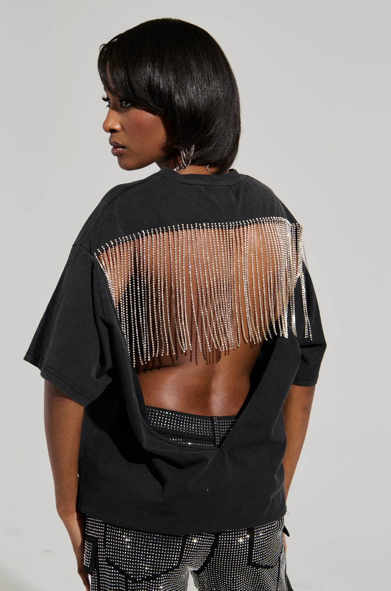A person with short black hair is wearing the SMOKE & MIRRORS RHINESTONE FRINGE MINERAL WASH T-SHIRT, featuring an open back and long, dangling rhinestone fringe. They pair it with sparkling pants and pose sideways against a neutral background. 4