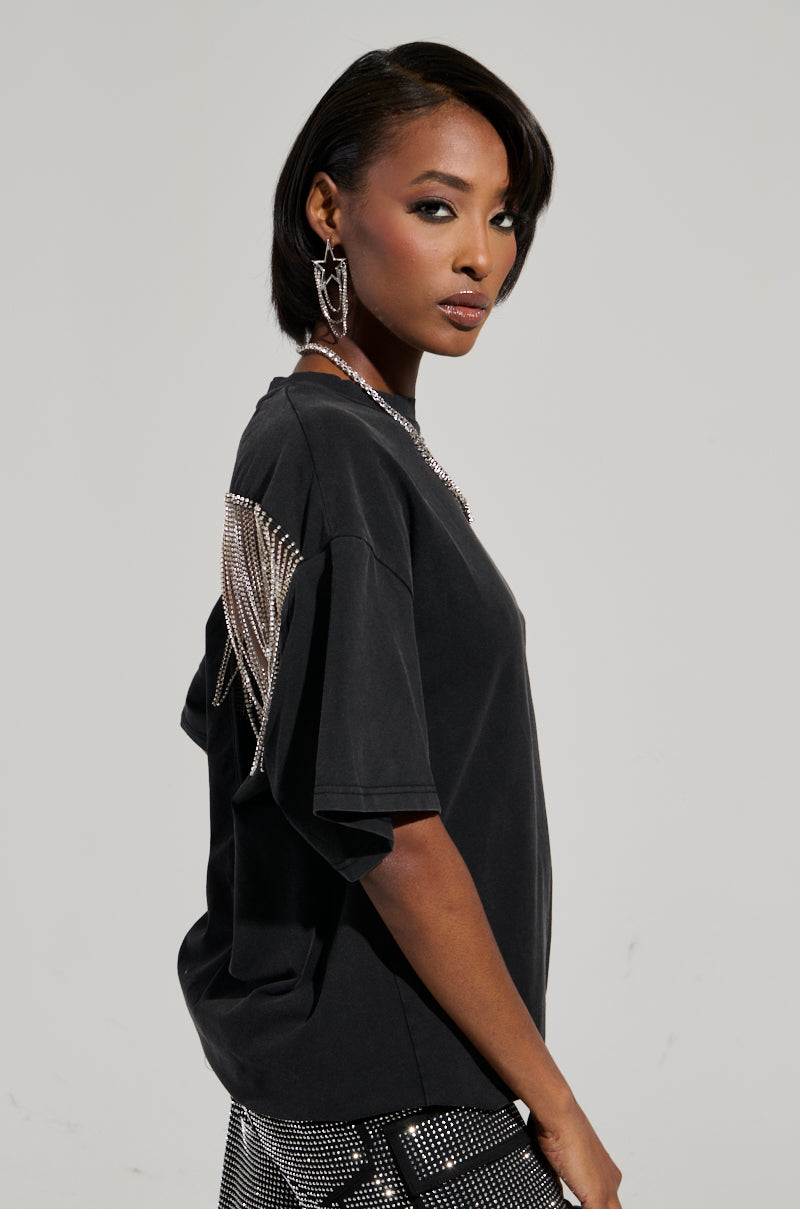 A woman with short hair wears the SMOKE & MIRRORS RHINESTONE FRINGE MINERAL WASH T-SHIRT in black, featuring sparkling embellishments on the back. She complements it with statement earrings and super stretchy matching pants, posing against a plain background and looking confidently at the camera. 5