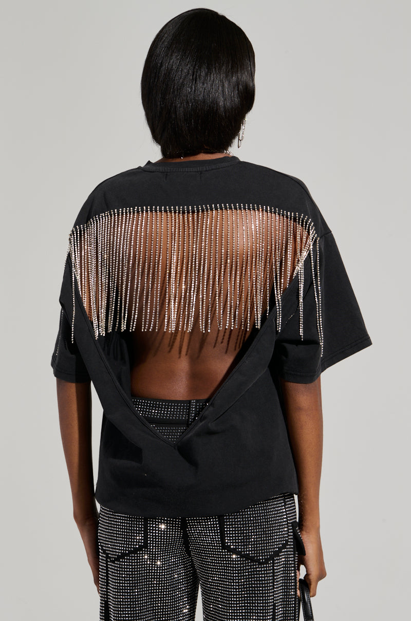 A person with short dark hair is viewed from behind, wearing the SMOKE & MIRRORS RHINESTONE FRINGE MINERAL WASH T-SHIRT, which features a cut-out back adorned with rhinestone fringe. Their studded pants enhance the edgy style, and they carry a small bag. The plain gray backdrop accentuates the ensemble's bold aesthetic. 6
