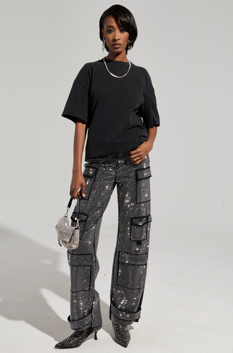 Wearing the SMOKE & MIRRORS RHINESTONE FRINGE MINERAL WASH T-SHIRT, a fashion model showcases her striking outfit, pairing it with glittery patterned pants that feature pockets. She carries a sparkling handbag and completes the look with matching sequined shoes, complemented by a necklace adorned with rhinestone fringe. The neutral background highlights her ensemble beautifully. 7