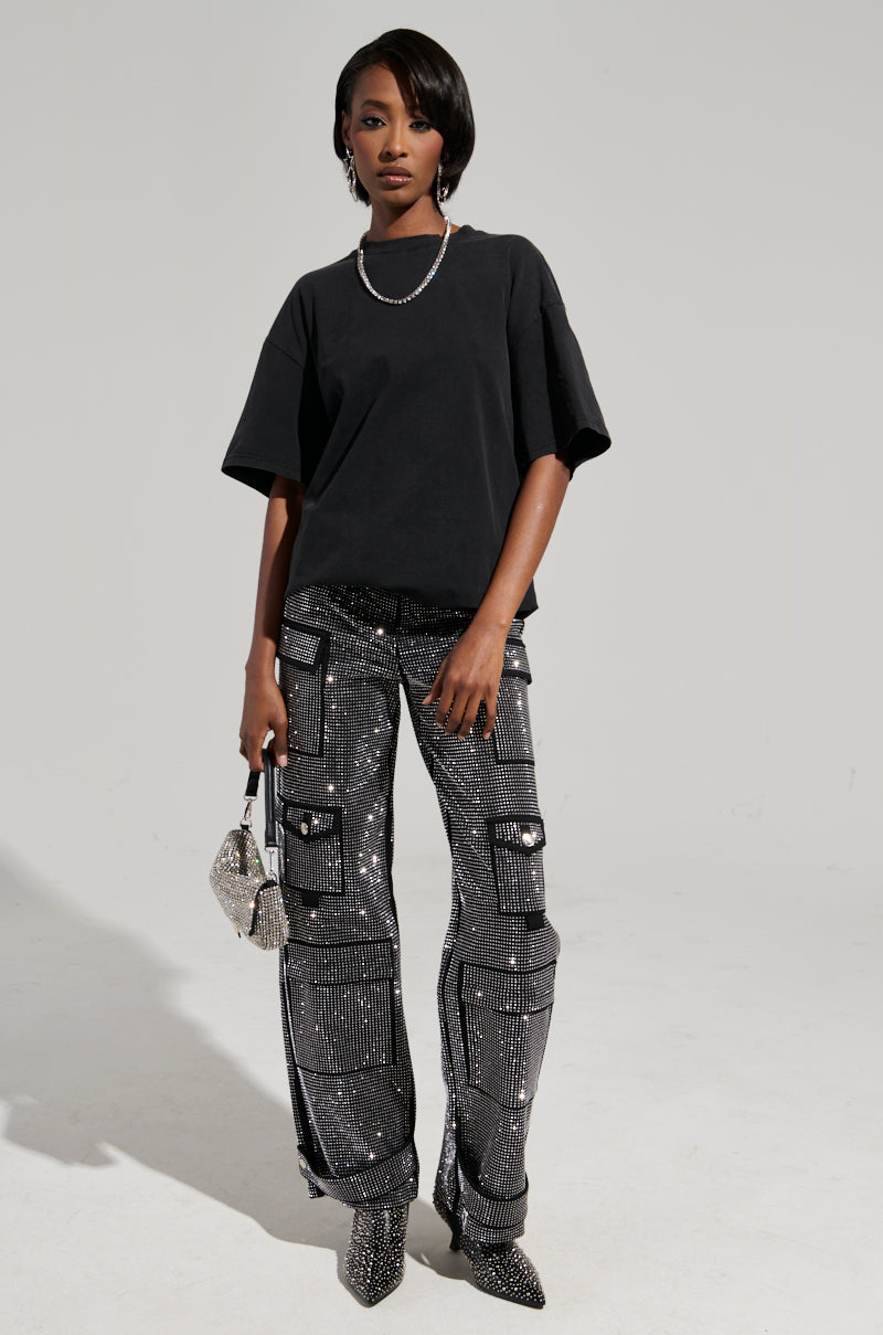 A person stands confidently wearing the Smoke & Mirrors Rhinestone Fringe Mineral Wash T-Shirt and shiny, sequined cargo pants. They hold a glittery purse with rhinestone fringe and wear sparkling pointed shoes. A silver necklace completes the stylish ensemble against a simple, uncluttered background. 8
