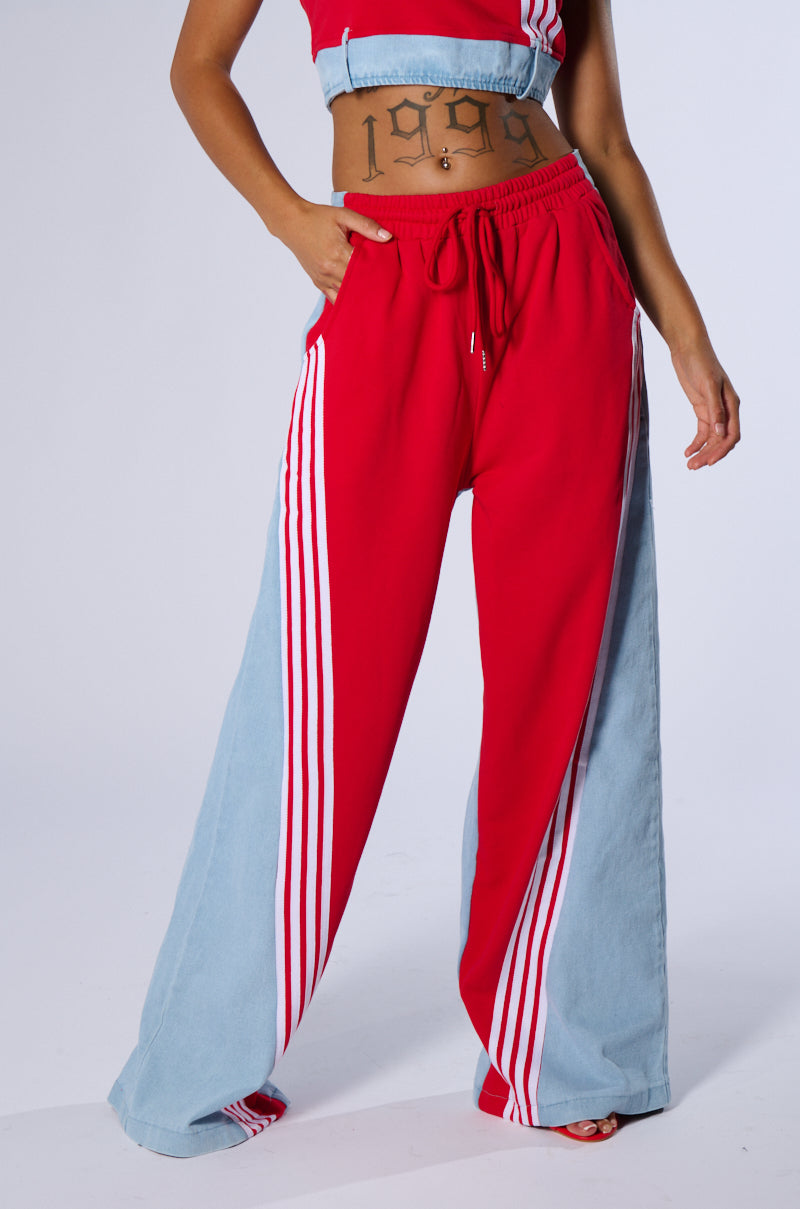 A person showcasing a sporty chic style is depicted wearing a red and blue athletic outfit. The high-waisted HEADS UP FLARE PANT are red with light blue panels on the sides, accented by white stripes. The matching crop top is also red with blue accents, perfectly coordinating with the pants. Notably, the individual has a "1999" tattoo across their stomach.