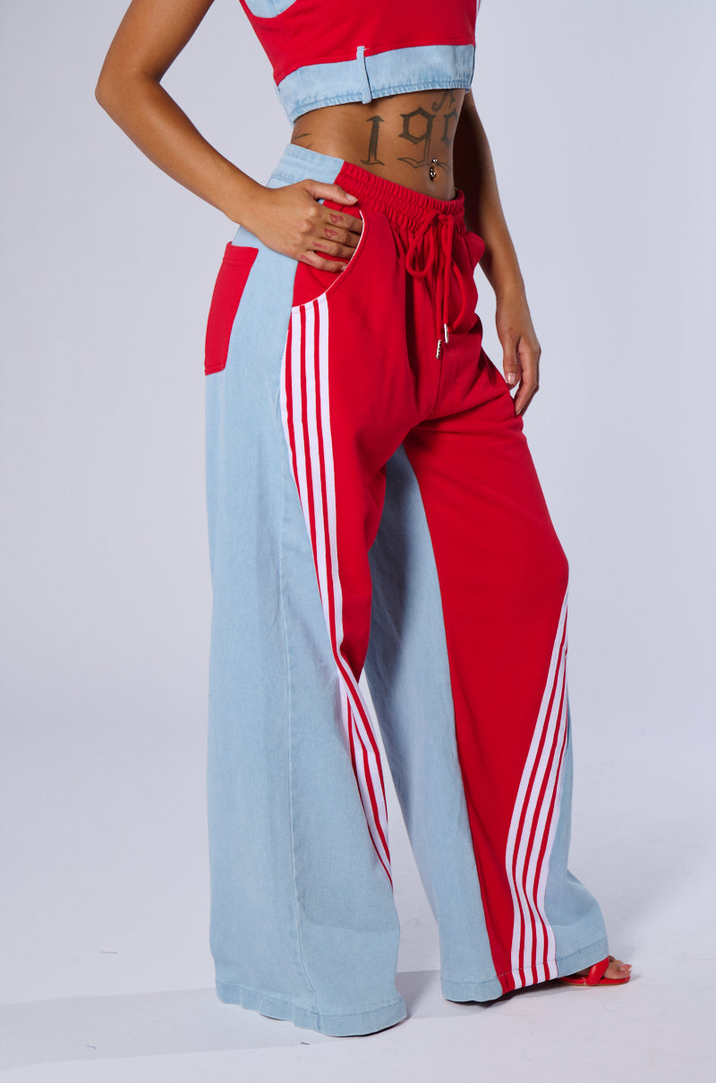 A person models the HEADS UP FLARE PANT, showcasing its high-rise design and wide-legged silhouette. The pants are a striking combination of bright red and light blue denim fabric, enhanced by vertical white side stripes that embody a sporty chic style. The individual completes the look with a matching cropped top, revealing some tattoos on their abdomen.