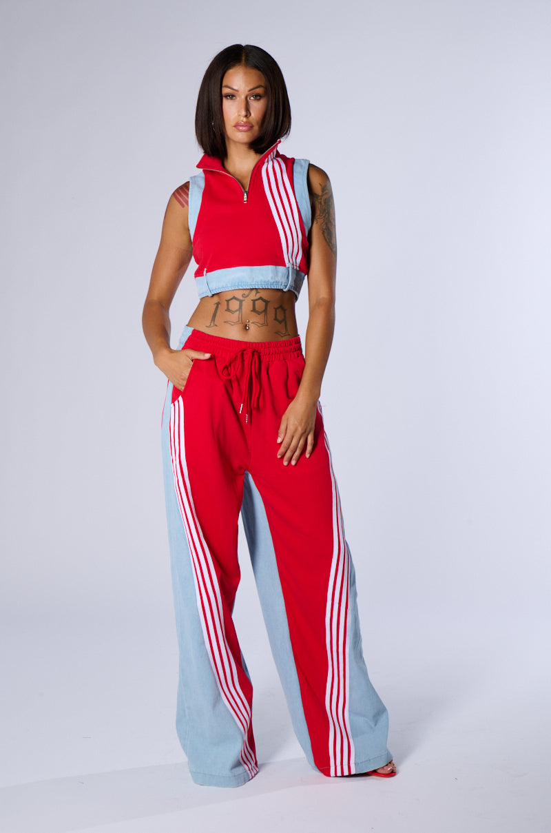 A woman stands confidently against a white background, wearing a sporty chic ensemble in matching red and light blue. The sleeveless top and Heads Up Flare Pants feature white vertical stripes. With her tattoo on display, hands in pockets, she flaunts her straight, mid-length hair.