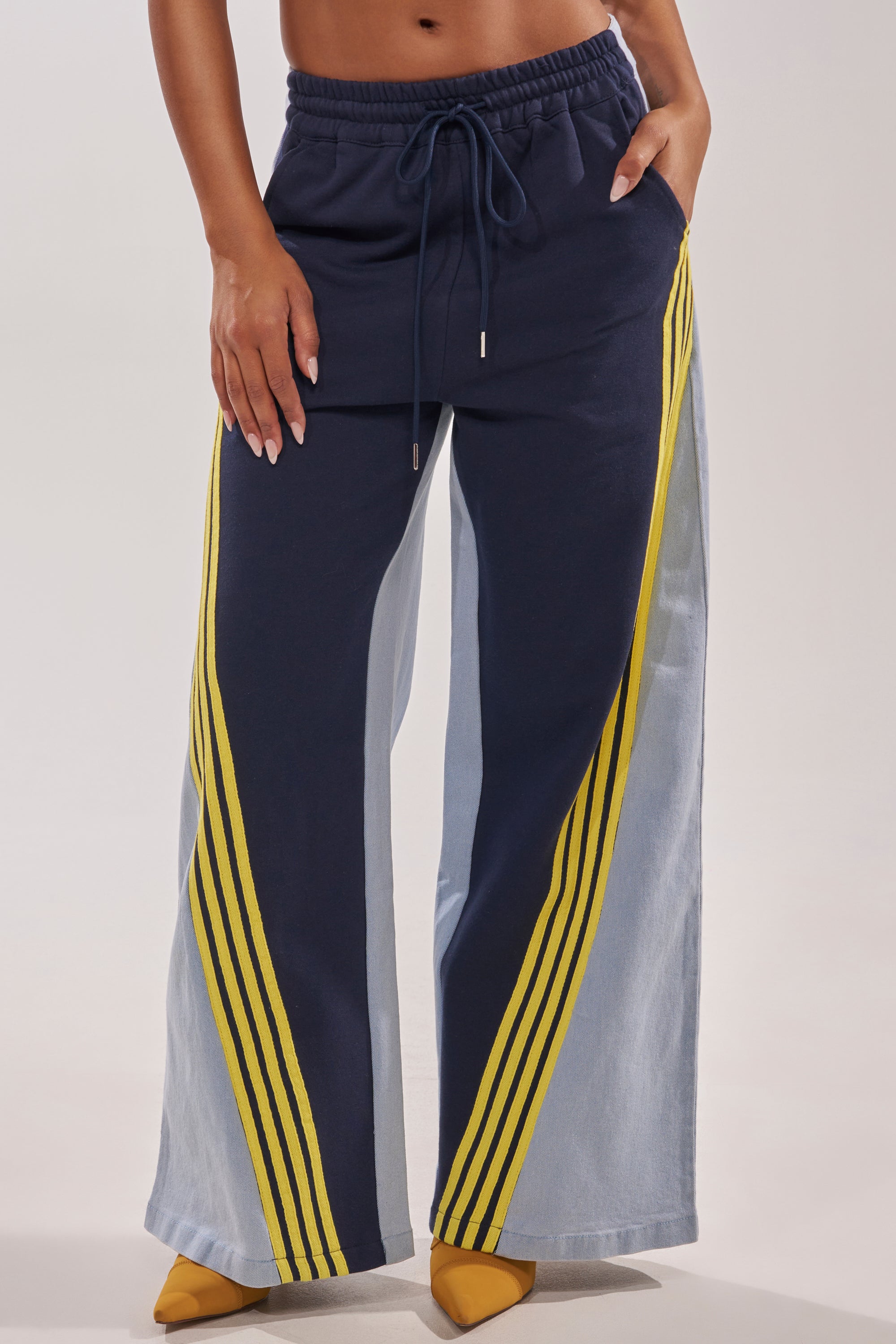 A person is wearing the HEADS UP DENIM WIDE LEG JOGGER IN NAVY, featuring yellow side stripes and light blue panels, paired with yellow pointed-toe heels. Only the lower half of the body is shown. 1