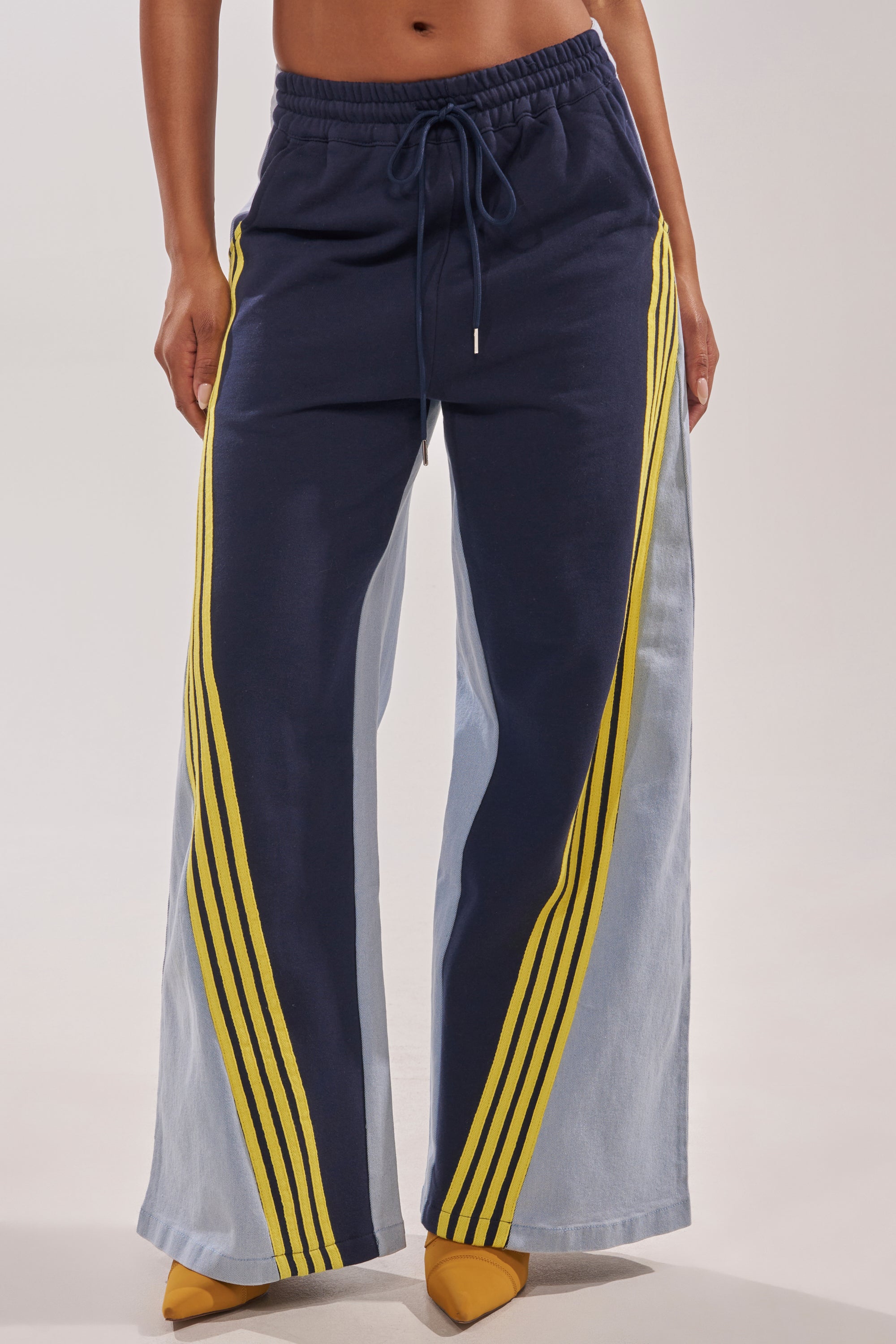 A person wears the HEADS UP DENIM WIDE LEG JOGGER IN NAVY featuring yellow stripes, light blue side panels, and a drawstring waist, paired with yellow pointed-toe shoes. The upper body is not visible. 2
