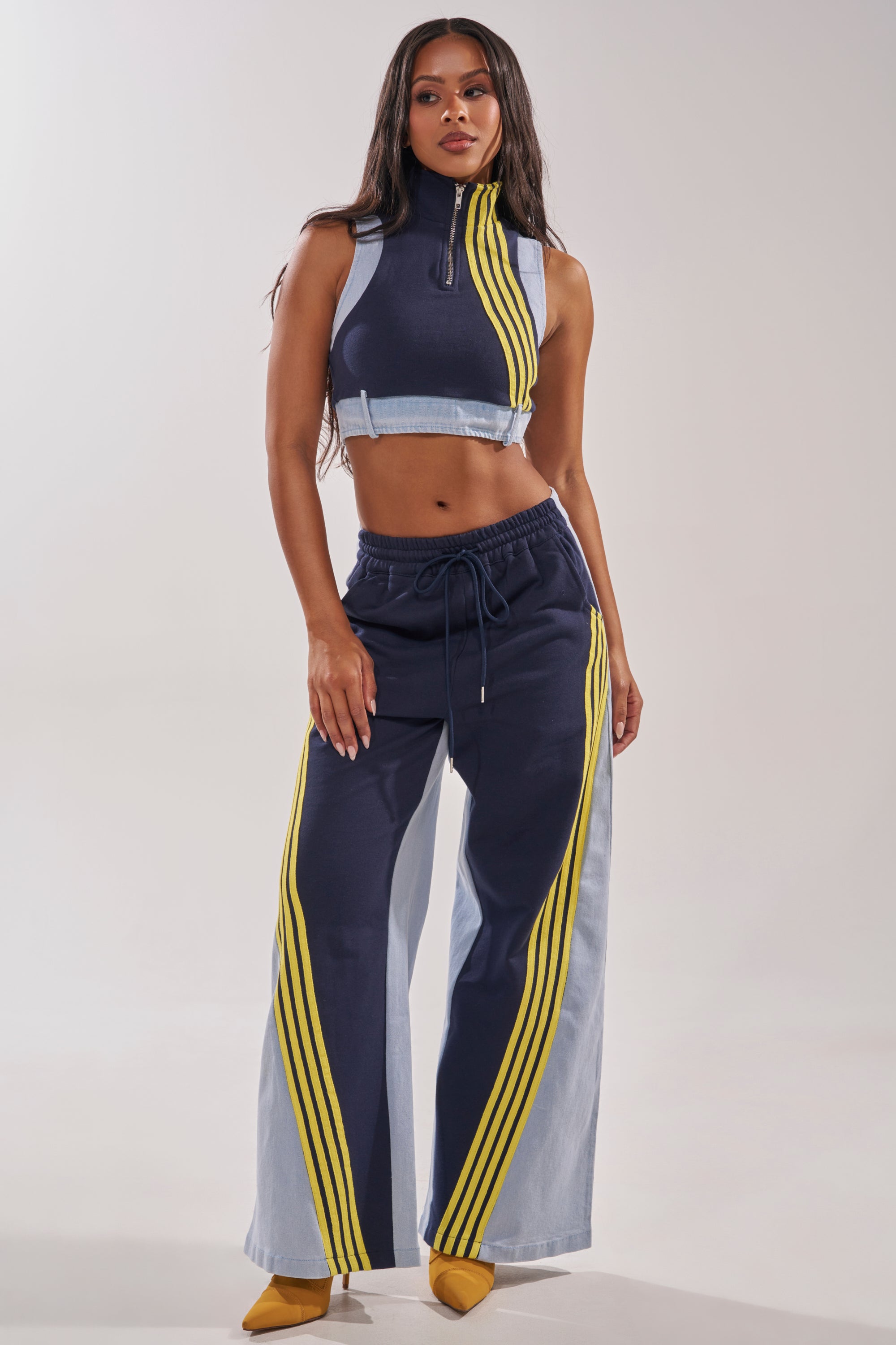 A woman stands confidently in the HEADS UP DENIM WIDE LEG JOGGER IN NAVY, styled with a navy crop top featuring yellow stripes and yellow pointed-toe heels. 5