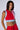 A woman with medium-length dark hair confidently poses in the HEADS UP TANK by AKIRA Label, a red and light blue sleeveless cropped top with a front zipper. Adopting a sporty style, she pairs it with red pants and proudly displays multiple tattoos, including the number "1993" on her stomach.