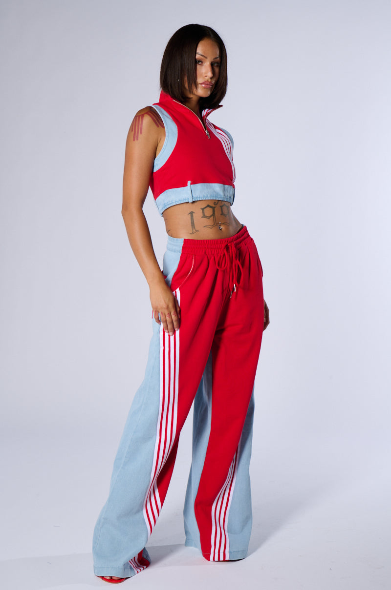 A person stands confidently, showcasing a sporty style with an AKIRA Label twist. They wear the HEADS UP TANK, a sleeveless, zip-up red crop top paired with matching red and light blue tracksuit pants featuring white stripes along the sides. The background is a simple, neutral grey.