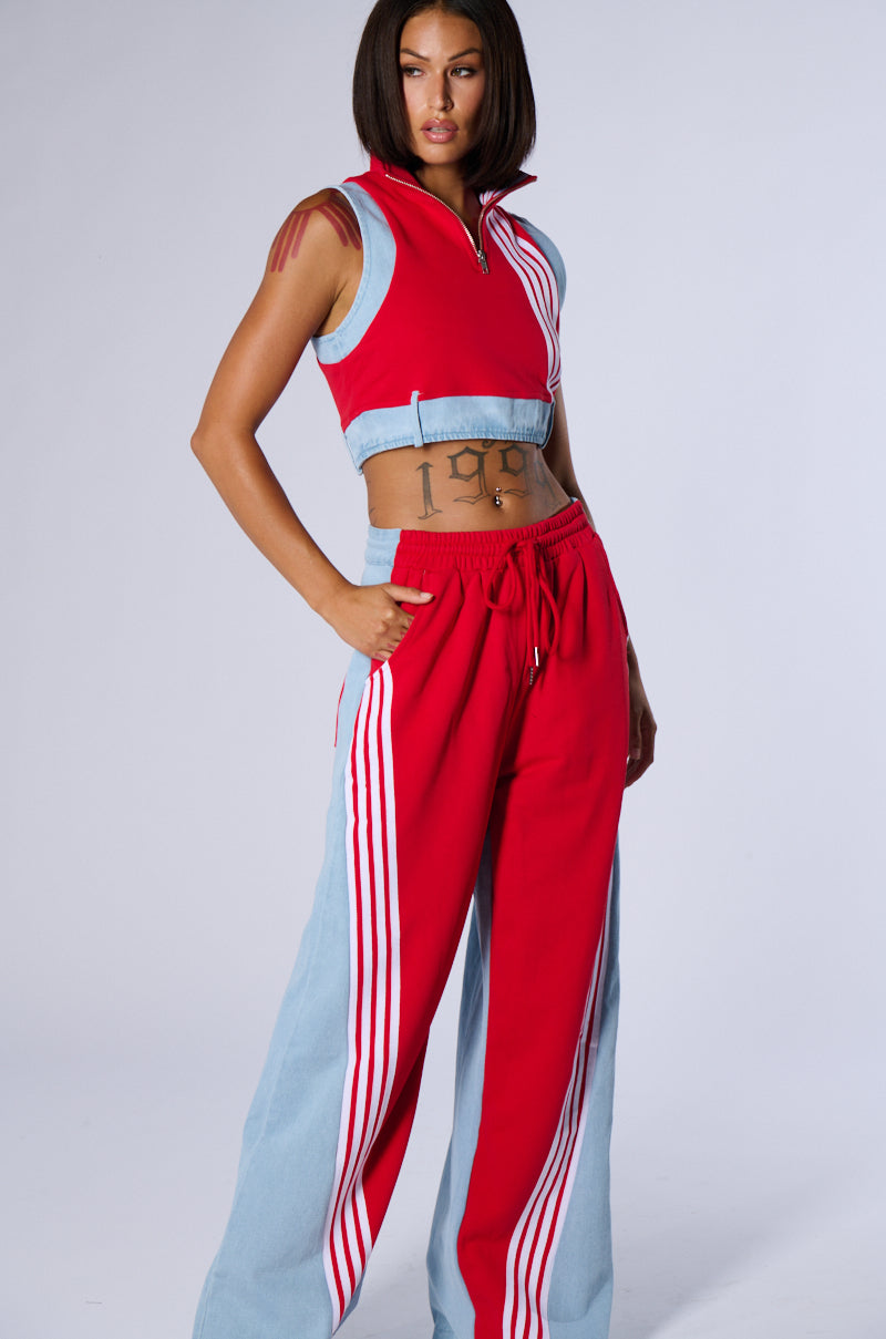 A person with short, dark hair poses confidently in a red and light blue tracksuit from the AKIRA Label, featuring the sleeveless HEADS UP TANK that reveals a tattoo of Roman numerals on their midriff. The pants boast red and white stripes against light blue panels, capturing a sporty style set against a plain light grey background.