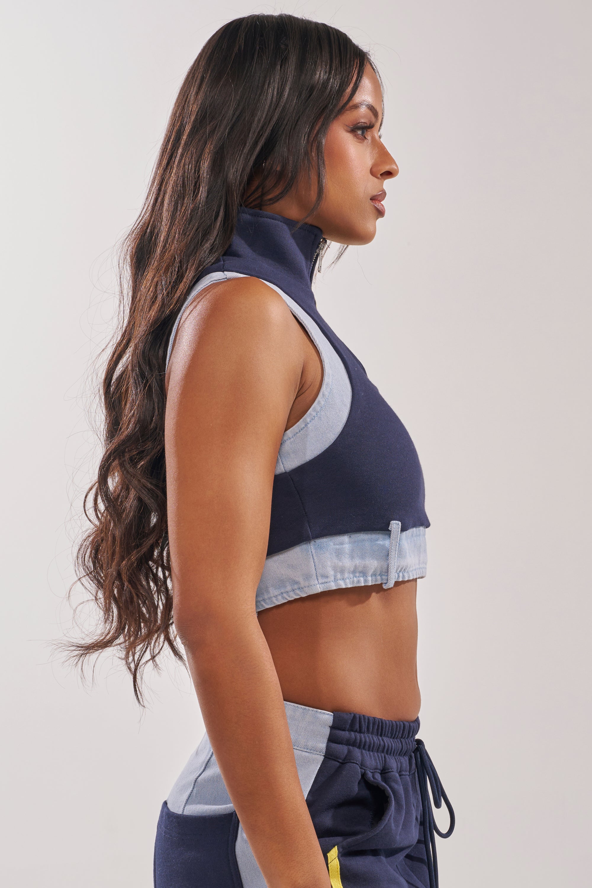 A woman with long wavy hair is shown in profile wearing the HEADS UP QUARTER ZIP SLEEVELESS TOP IN NAVY with matching drawstring pants and denim trim, standing against a plain light background. 4