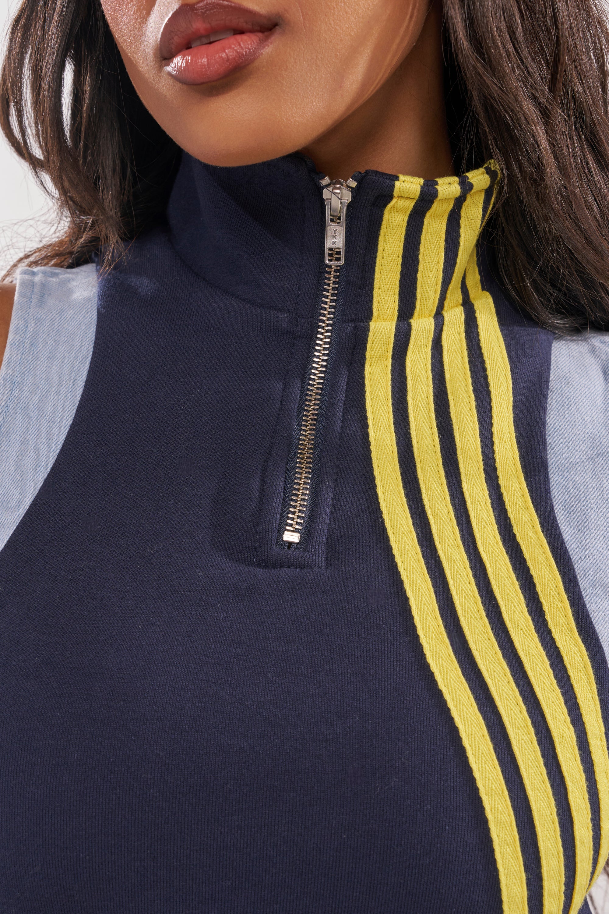 A close-up of a woman wearing the HEADS UP QUARTER ZIP SLEEVELESS TOP IN NAVY, featuring sporty-chic style with three yellow stripes on one side. Only her lower face, neck, and upper chest are visible. 7