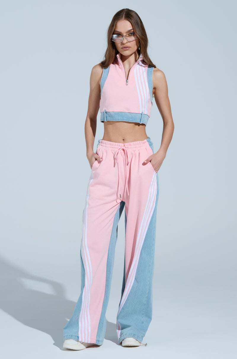 In a chic ensemble, a person sports the "HEADS UP QUARTER ZIP SLEEVELESS SWEATSHIRT IN PINK" matched with wide-legged pants accented with athletic stripes. They have long hair, wear glasses, and pose confidently against a plain background.