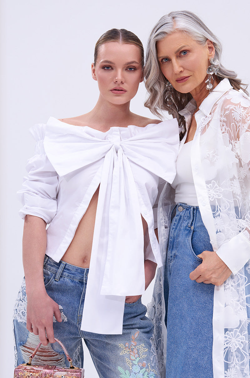 Two women pose together. One wears the BOWED UP BLOUSE featuring an oversized bow, paired with embroidered jeans. The other, with long gray hair, sports a white tank top, sheer floral coat, and blue jeans.