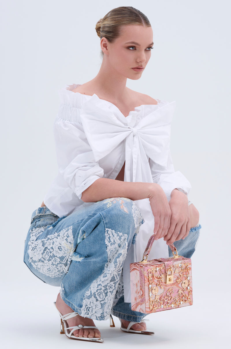 A woman squats wearing the BOWED UP BLOUSE in white poplin with blue jeans featuring white lace patches, white heels, and a pink floral handbag. Her hair is styled in a neat bun.