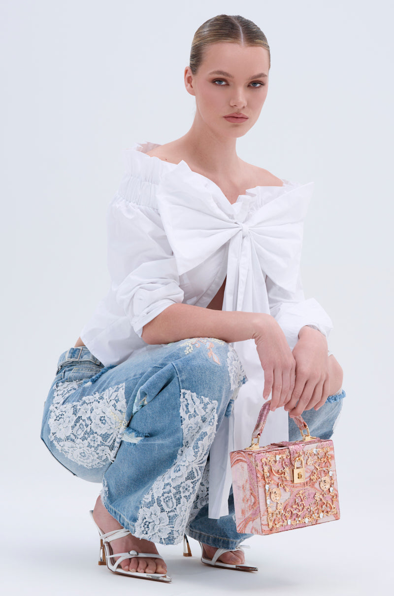 A woman with slicked-back hair squats, wearing the BOWED UP BLOUSE with off-shoulder poplin details, blue jeans with white lace panels, white strappy heels, and holding a small ornate pink and gold handbag.