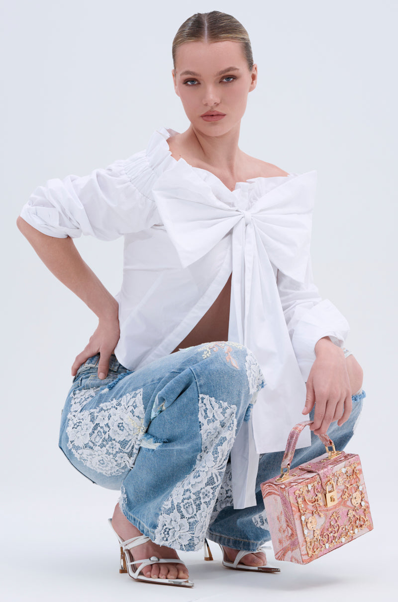 A woman squats confidently, wearing the BOWED UP BLOUSE with an off-shoulder white bow design, blue jeans with lace patches, white heeled sandals, and holding a pink ornate handbag. Her hair is sleek and styled.