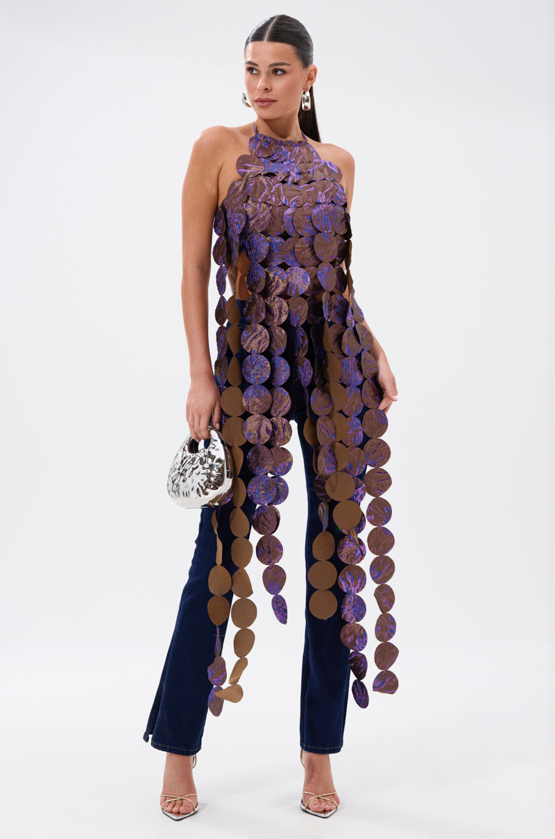 A woman wears the LIGHTS FLASHING BROCADE HALTER TIE NECK, featuring sequin appliqué with large brown discs, paired with dark blue pants. She holds a small silver purse, wears heels, and has her hair pulled back sleekly. 1