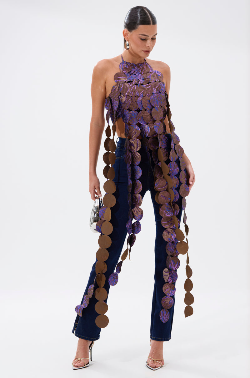 A woman poses in flared navy jeans, silver heels, and the LIGHTS FLASHING BROCADE HALTER TIE NECK top made of brown and purple sequin discs. She carries a small silver purse and wears her hair slicked back. 2
