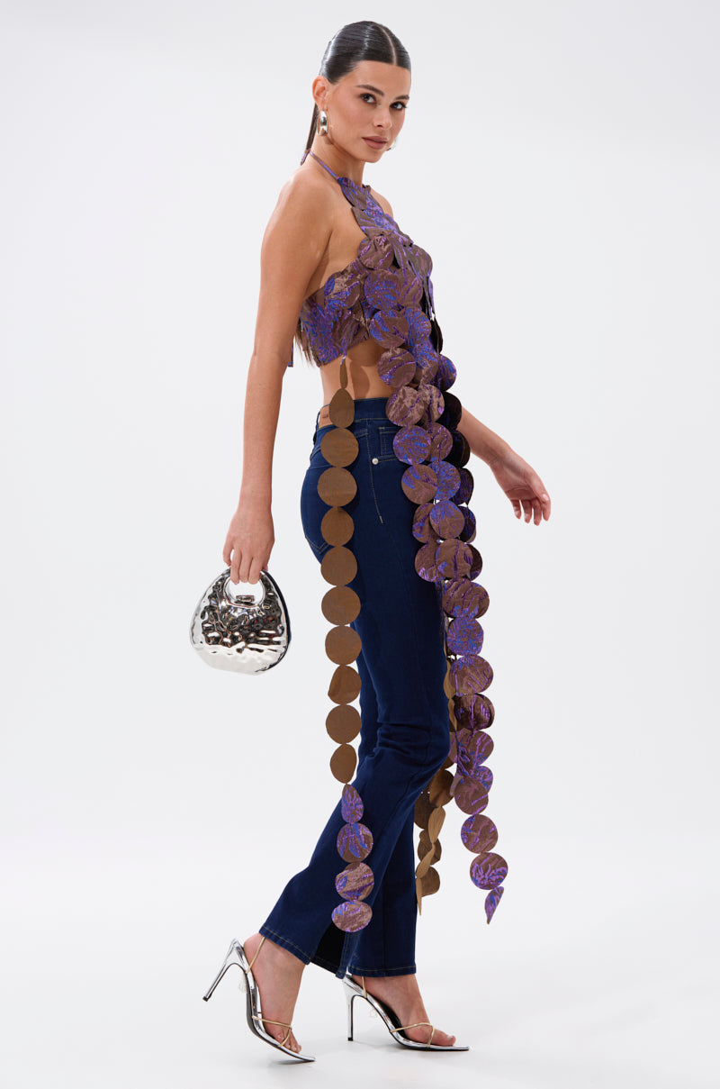 A woman poses confidently against a plain white background wearing the LIGHTS FLASHING BROCADE HALTER TIE NECK, dark blue jeans, silver high-heeled sandals, and holding a silver metallic handbag. 3
