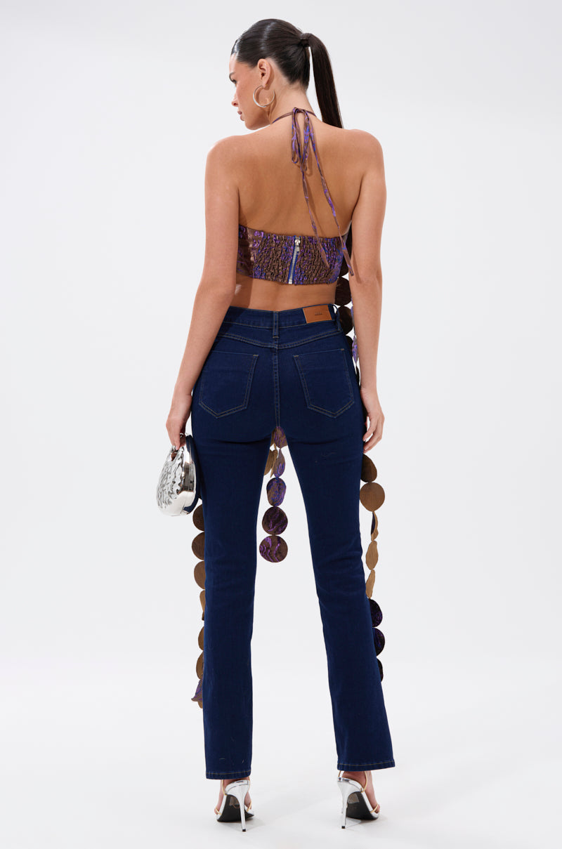 A woman with long dark hair in a ponytail faces away, wearing the LIGHTS FLASHING BROCADE HALTER TIE NECK, dark blue high-waisted jeans, high-heeled sandals, and holding a small silver bag against a plain white backdrop. 4