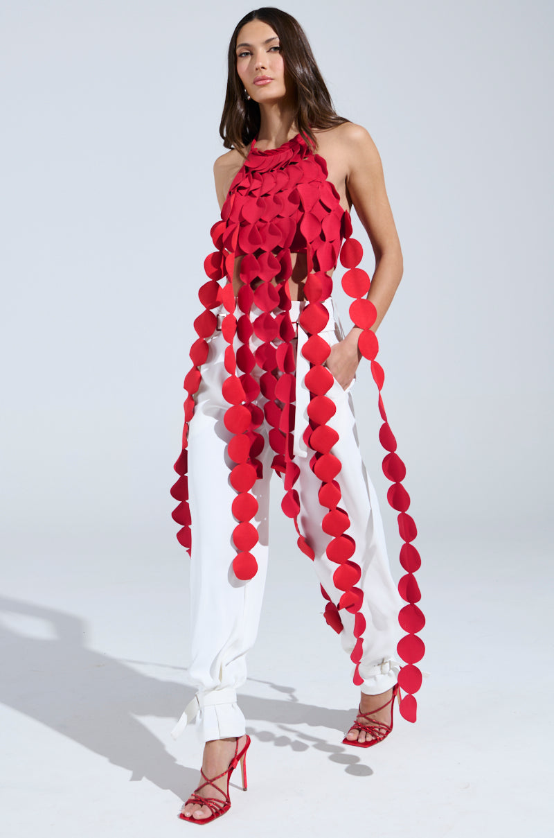 A woman models the LIGHTS FLASHING HALTER TIE NECK TOP IN RED, featuring large circular fabric pieces hanging in long strands. She pairs it with white pants and red heeled sandals against a plain light background.
