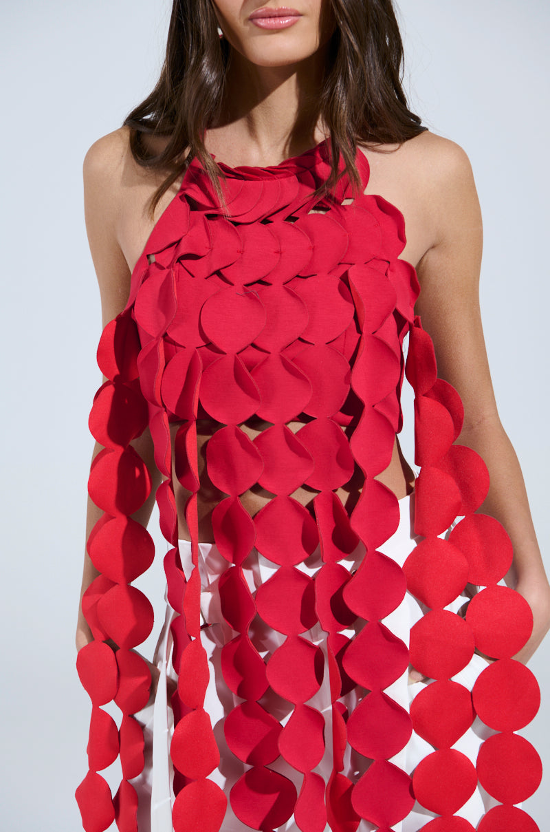 A woman wears the LIGHTS FLASHING HALTER TIE NECK TOP IN RED, featuring large overlapping circular pieces and long hanging strips, paired with white pants. Her face is partially visible and her hair is worn loose.